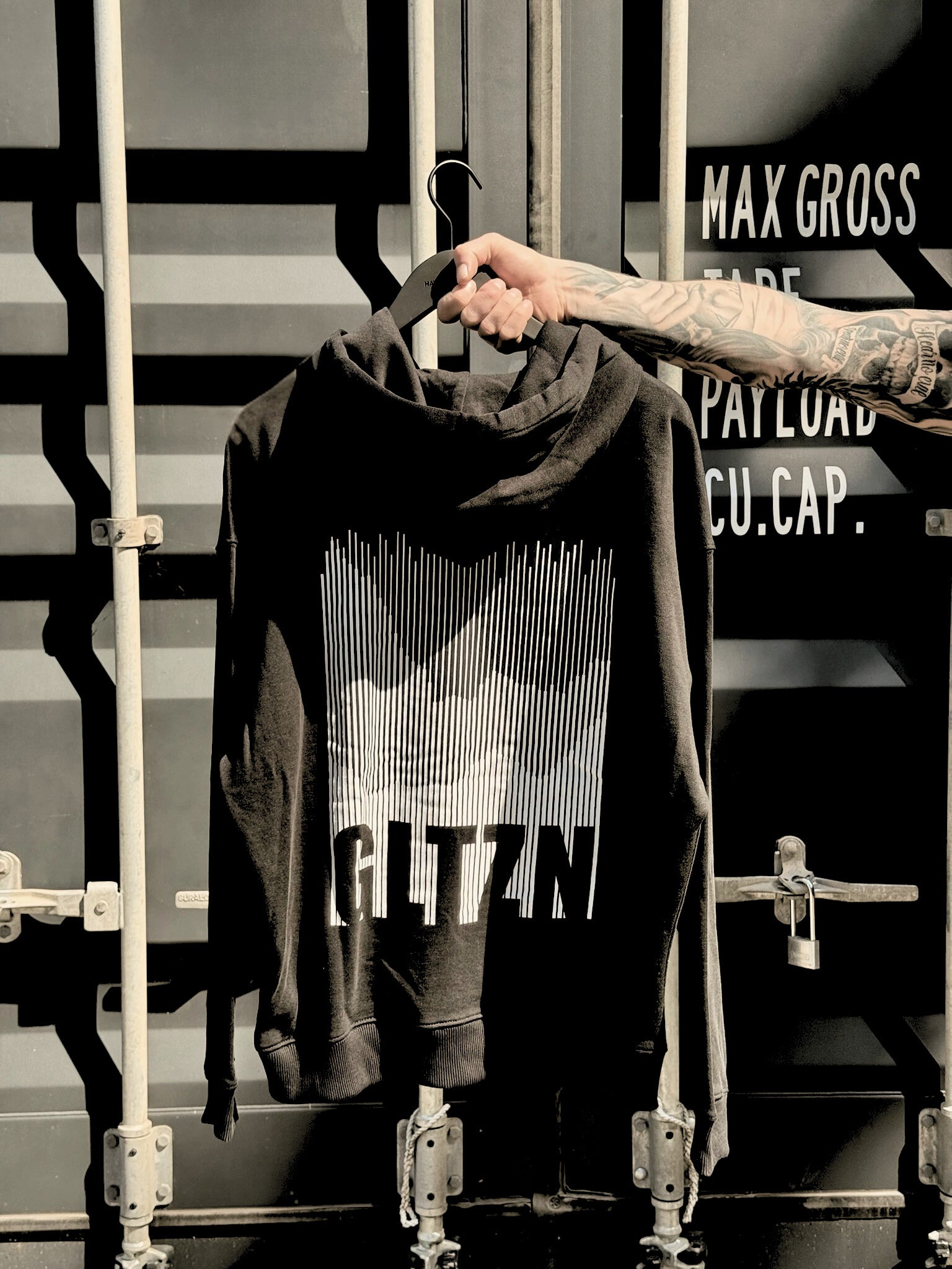 Ein tattowierter Männerarm halt die Rückseite eines schwarzen GLTZN Hoodie mit weißem Mountainprint vor einem dunkelgraunen Container