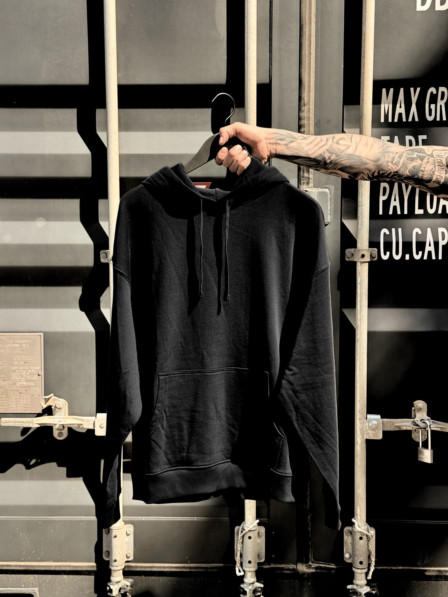 Ein tattowierter Männerarm halt die Vorderseite eines schwarzen GLTZN Hoodie vor einem dunkelgraunen Container