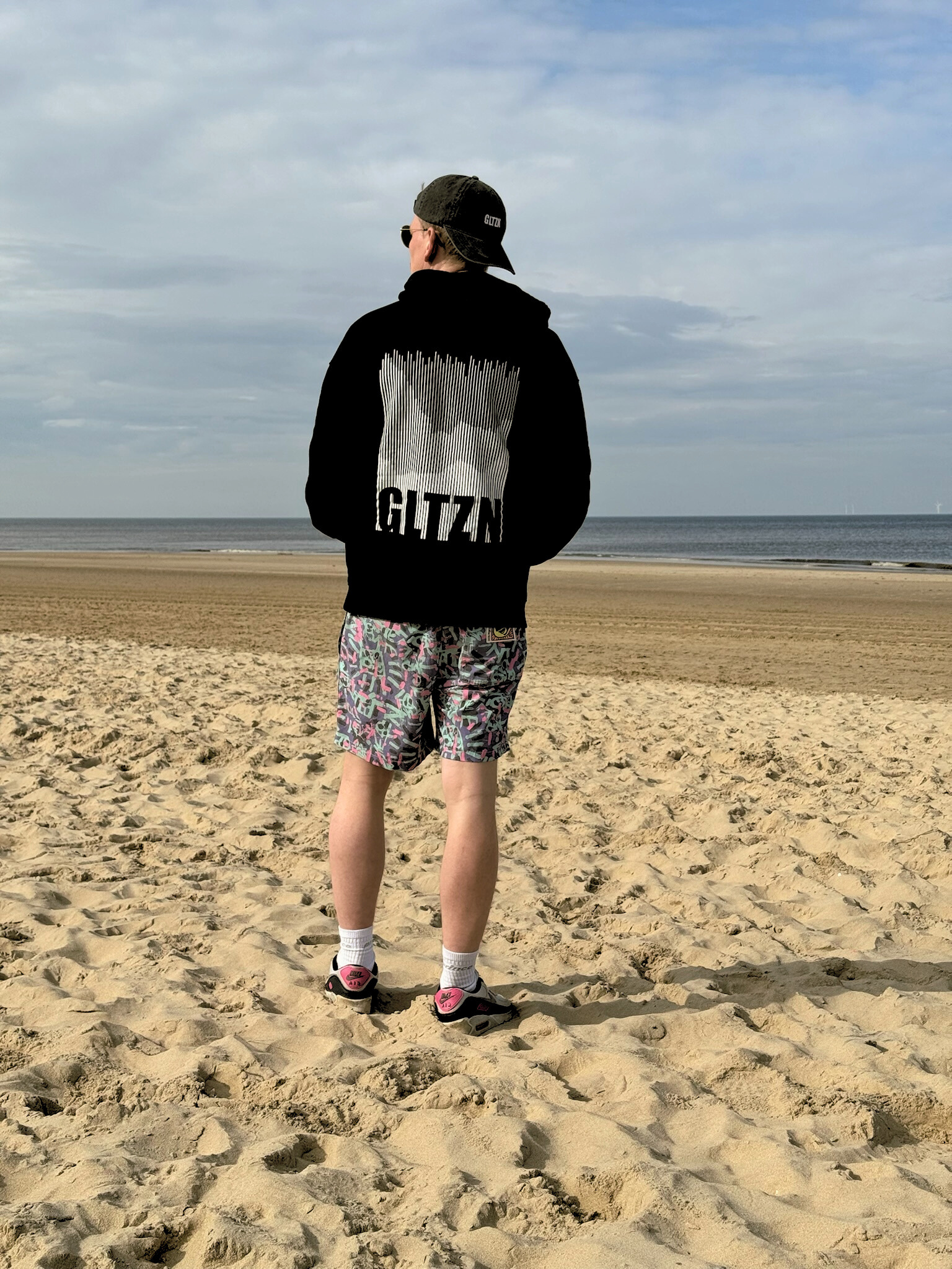 Mann mit Kappe steht mit schwarzen GLTZN-Hoodie mit Mountainprint und in Badehose mit dem Rücken zur Kamera am Meer