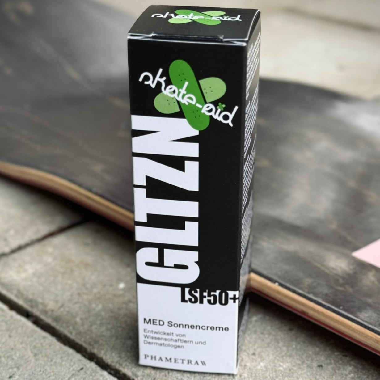 Die schwarz weiße Umverpackung von GLTZN Med Sonnencreme mit Skate Aid-Logo steht neben einem Skateboard auf Pflastersteinen