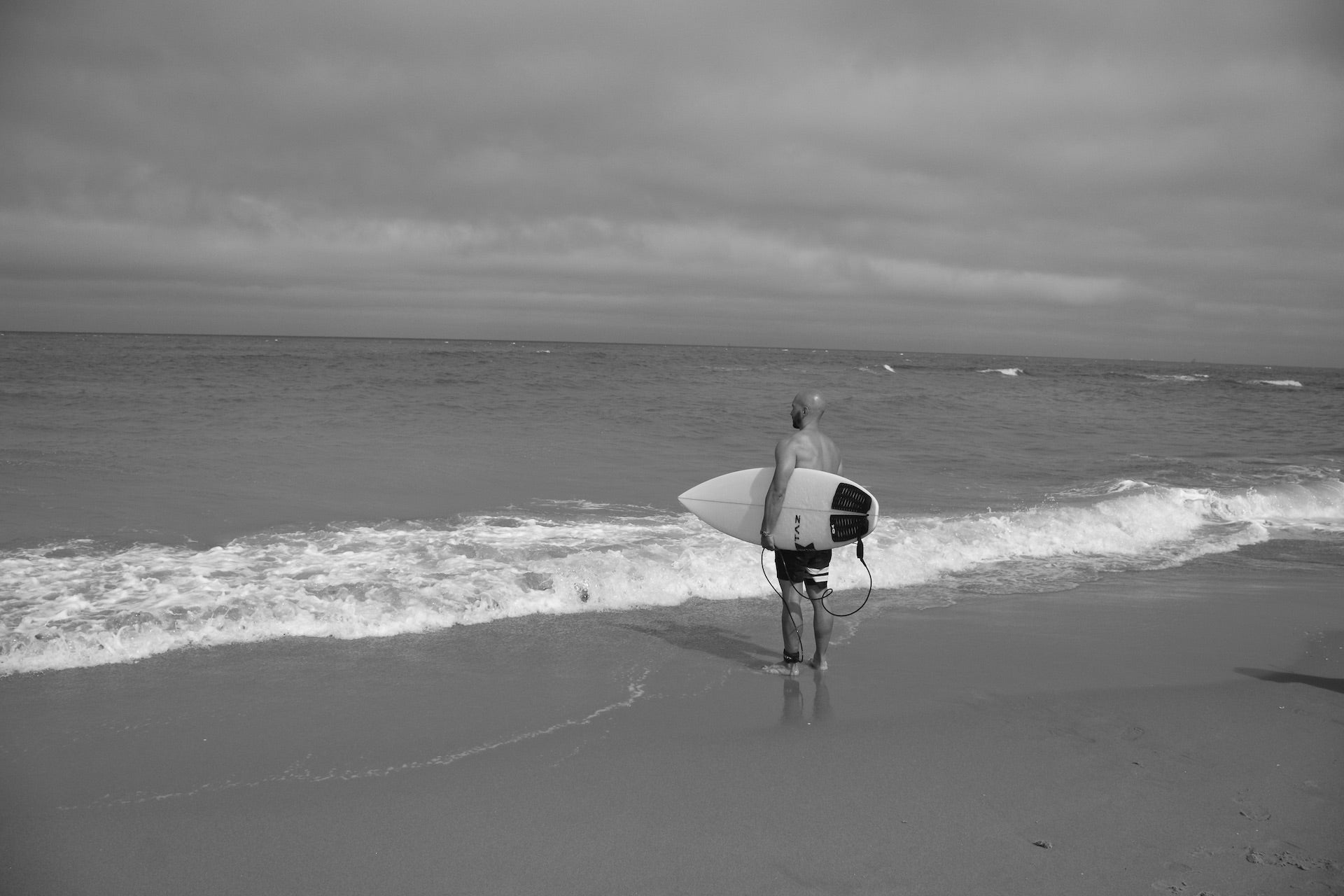 Surfer mit Glatze steht am Meer und hält Surfboard im Arm