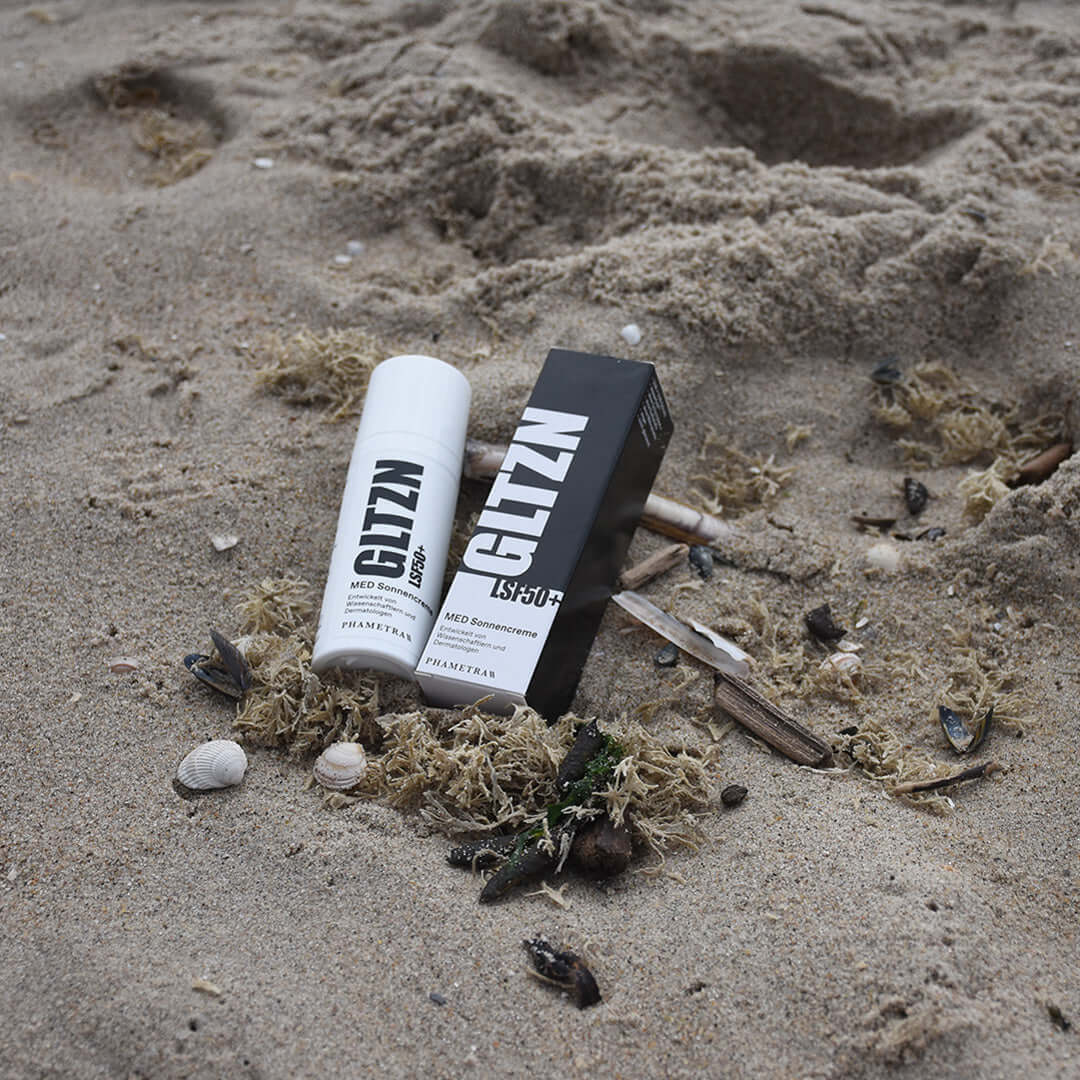 Ein weißer Spender GLTZN Med Sonnencreme in schwarzer Schrift und die schwarz-weiße Umverpackung liegen nebeneinander im Sand, umgeben von Moos, Muscheln und Steinchen