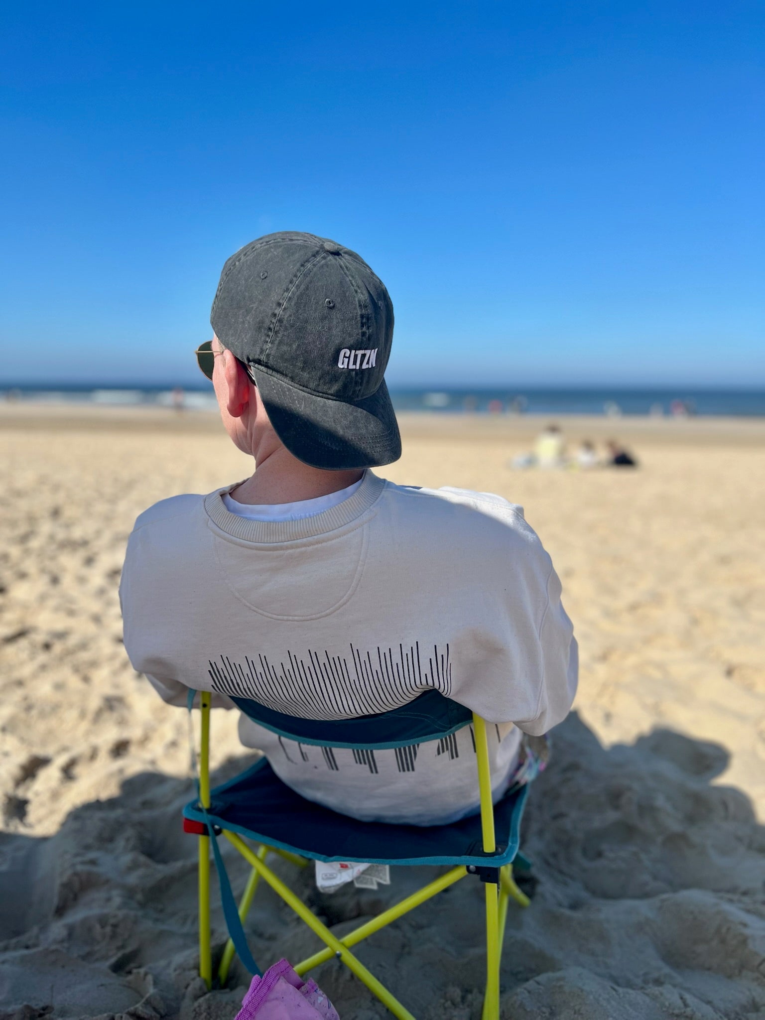 Mann sitzt am Strand auf einem neongelben Klappstuhl und trägt einen beigen Pulli sowie eine Sonnebrille und eine graue Kappe mit GLTZN Print. Er wird von hinten gezeigt.