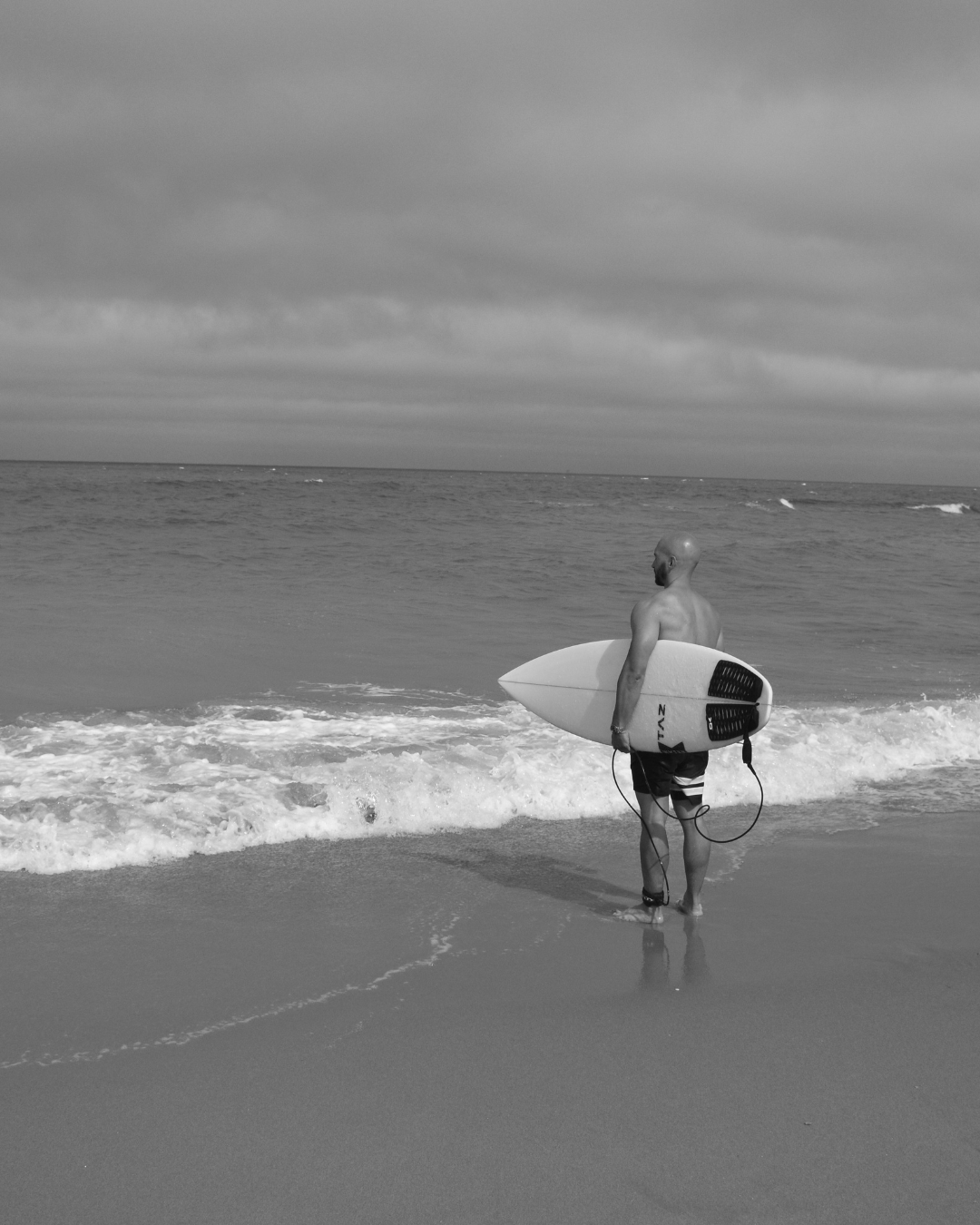 Mann mit Glatze hält Surfboard und steht vor Meer.