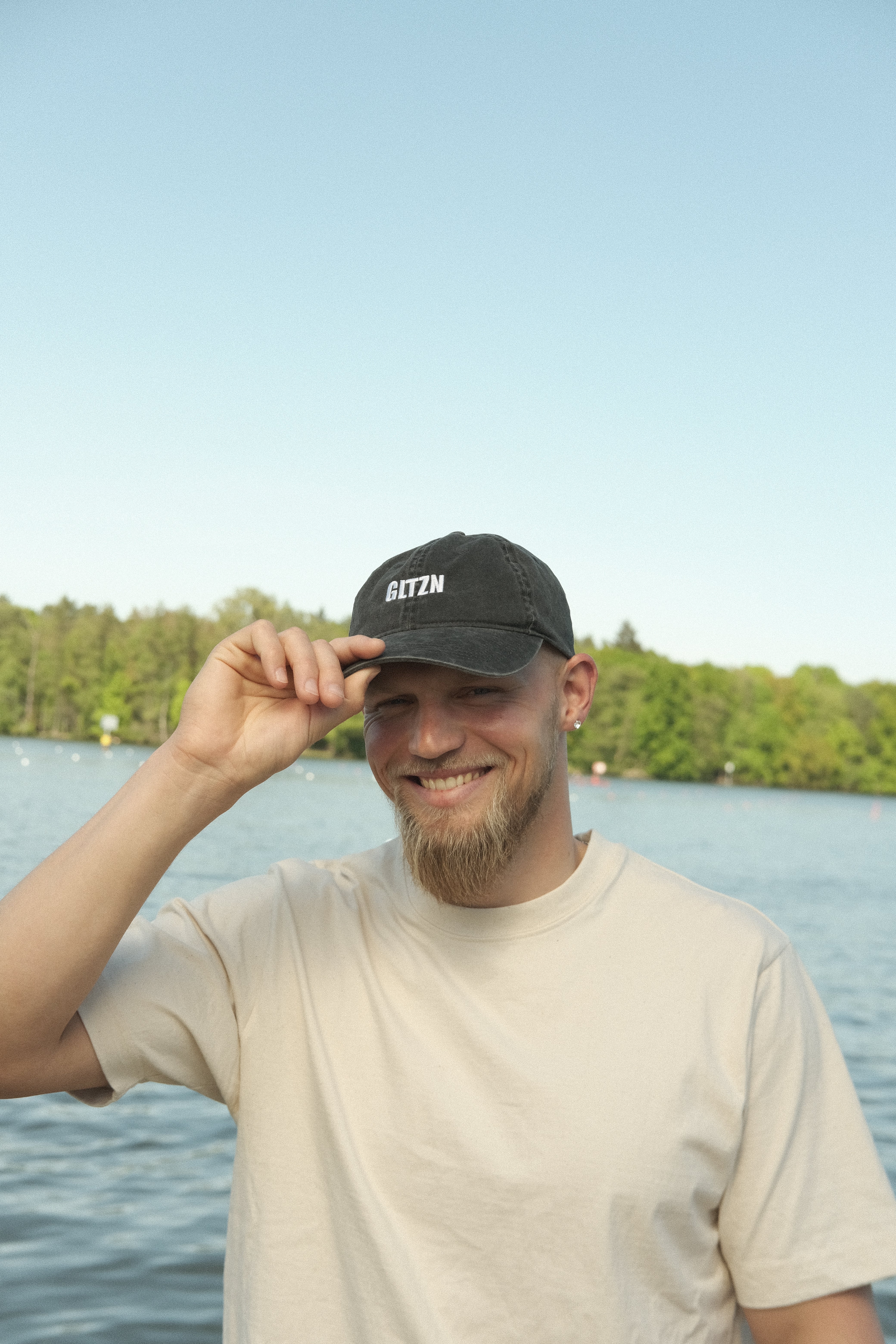 Ein Mann mit Bart und beigem T-Shirt trägt eine graue Cap mit GLTZN-Print und fasst an die Krempe der Kappe