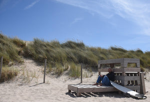 Mann mit Glatze sitzt in den Dünen neben Surfboard