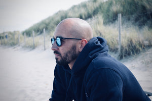 Mann mit Glatze und Sonnenbrille sitzt am Strand mit Dünen im Hintergrund