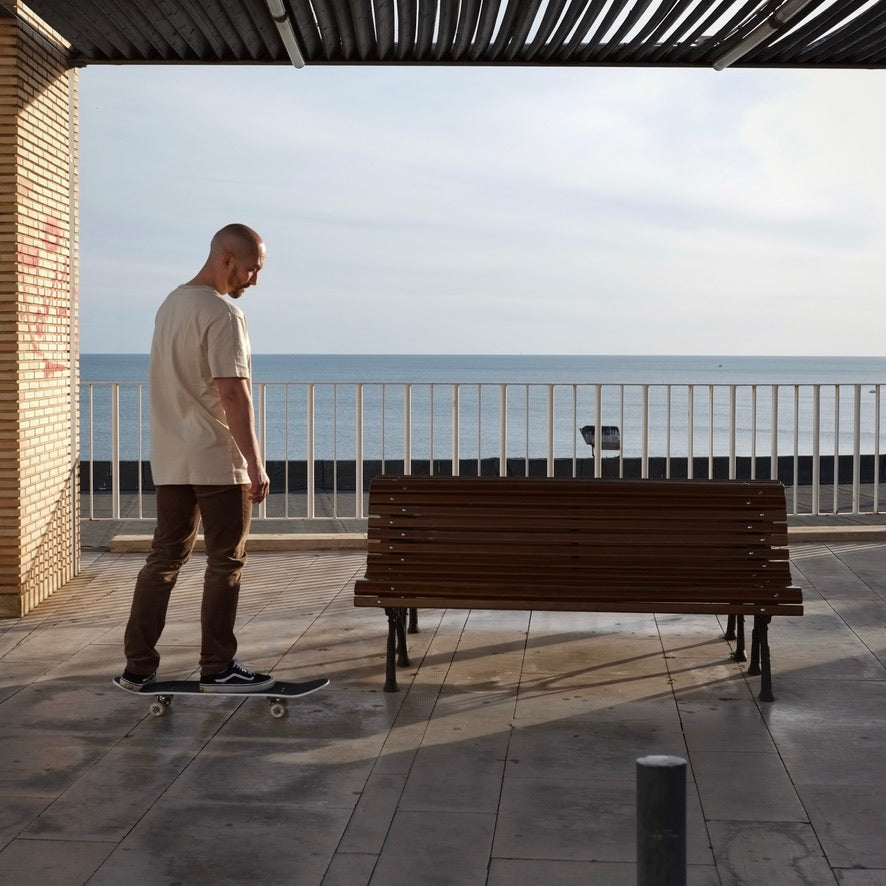 Ein Mann mit Glatze steht auf einem Skateboard vor einer Parkbank, im Hintergrund ist das Meer zu sehen
