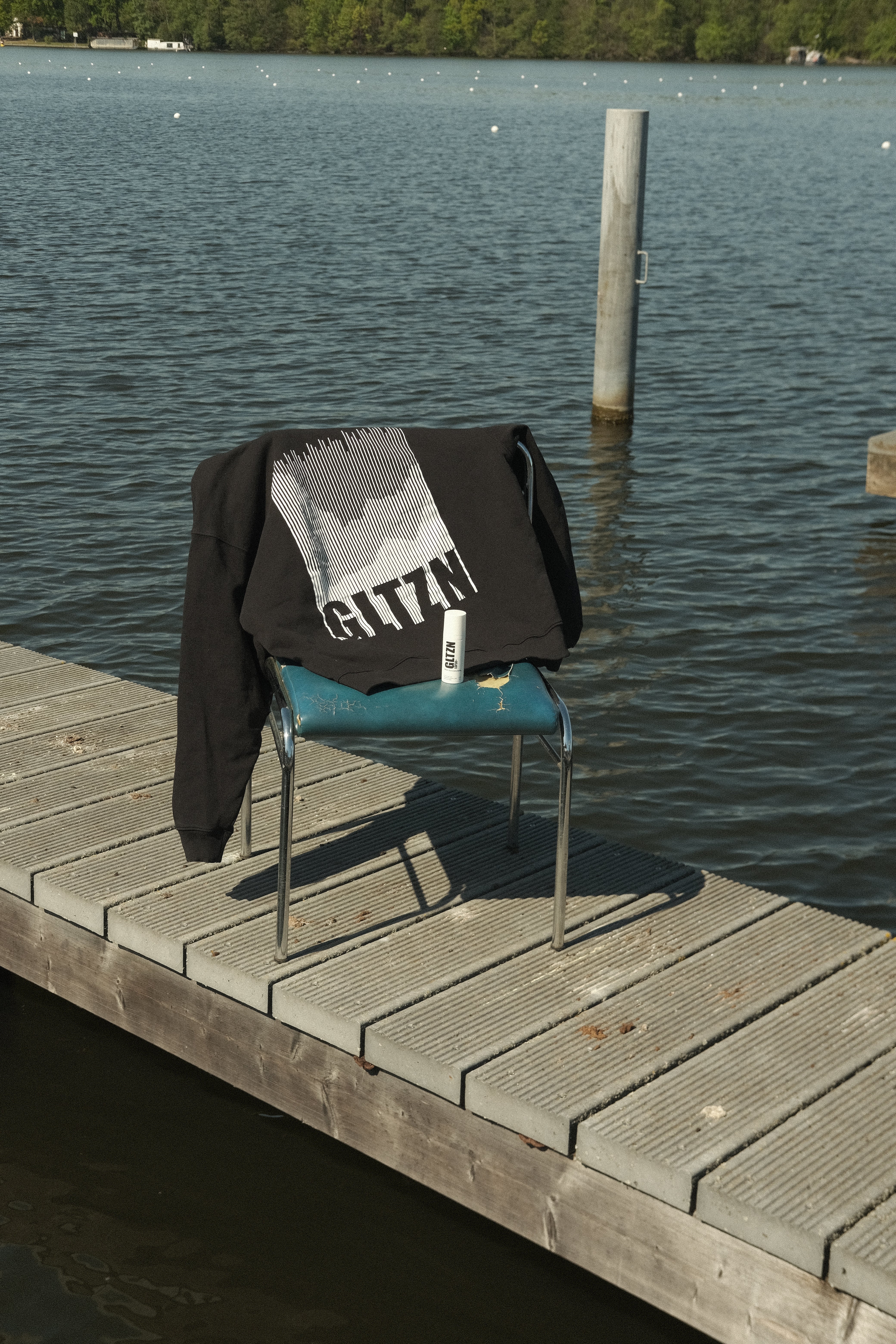Ein schwarzer GLTZN-Hoodie mit Mountainprint liegt auf einem kaputten, blauen Stuhl neben der GLTZN Sonnencreme auf einem Steg aus Holz