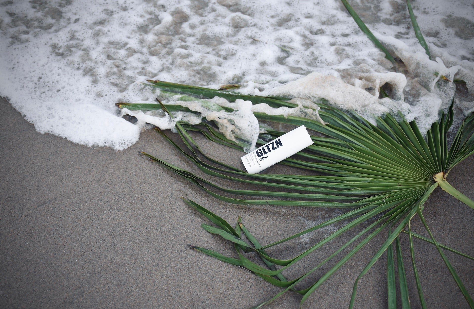 Ein schwarz weißer Spender mit GLTZN med Sonnencreme liegt am Strand auf einem Palmenplatt und wird vom Meer umspült