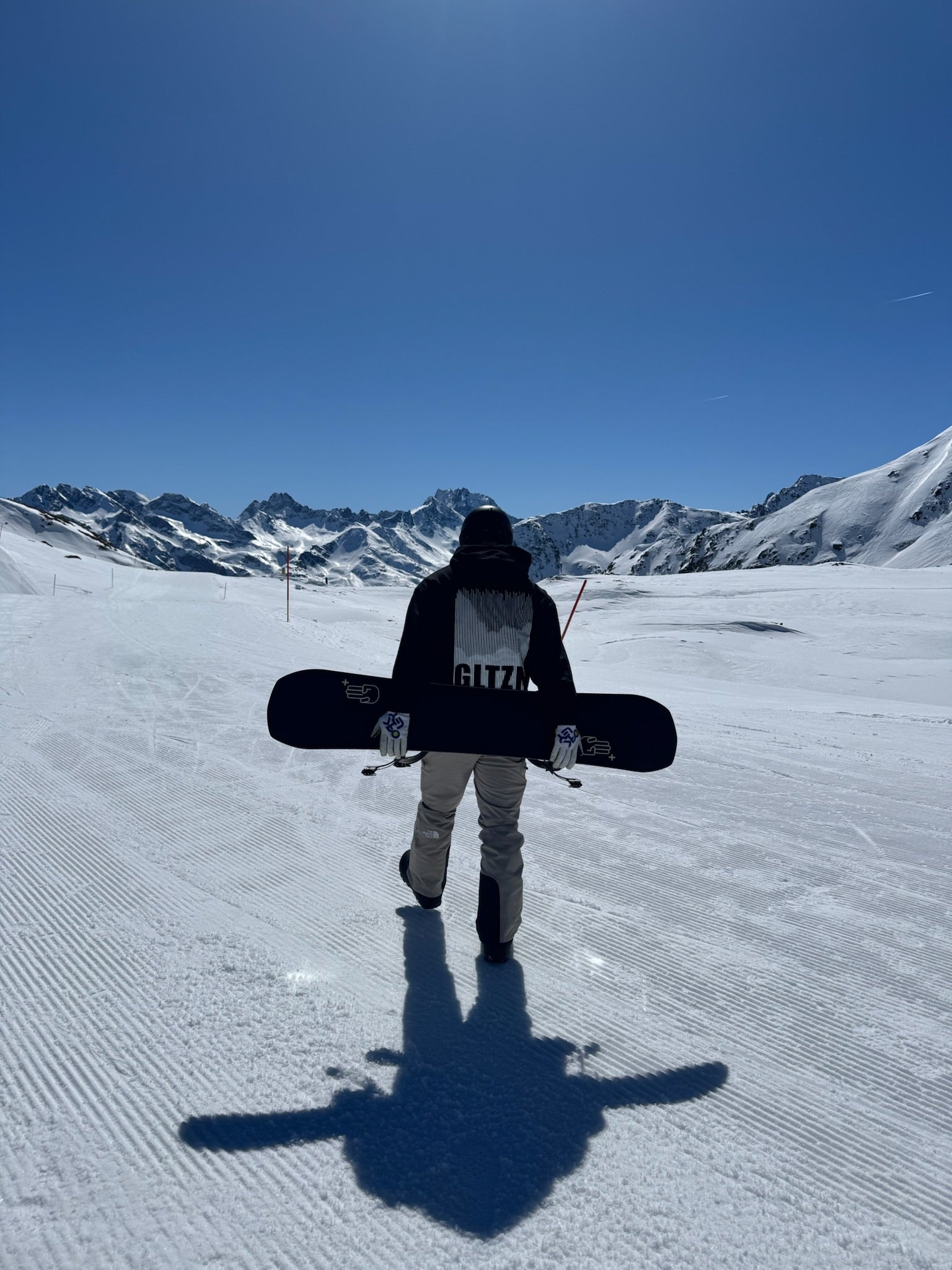 Snowboarder mit schwarzem Pulli und der Aufschrift "GLTZN" steht auf der Piste.