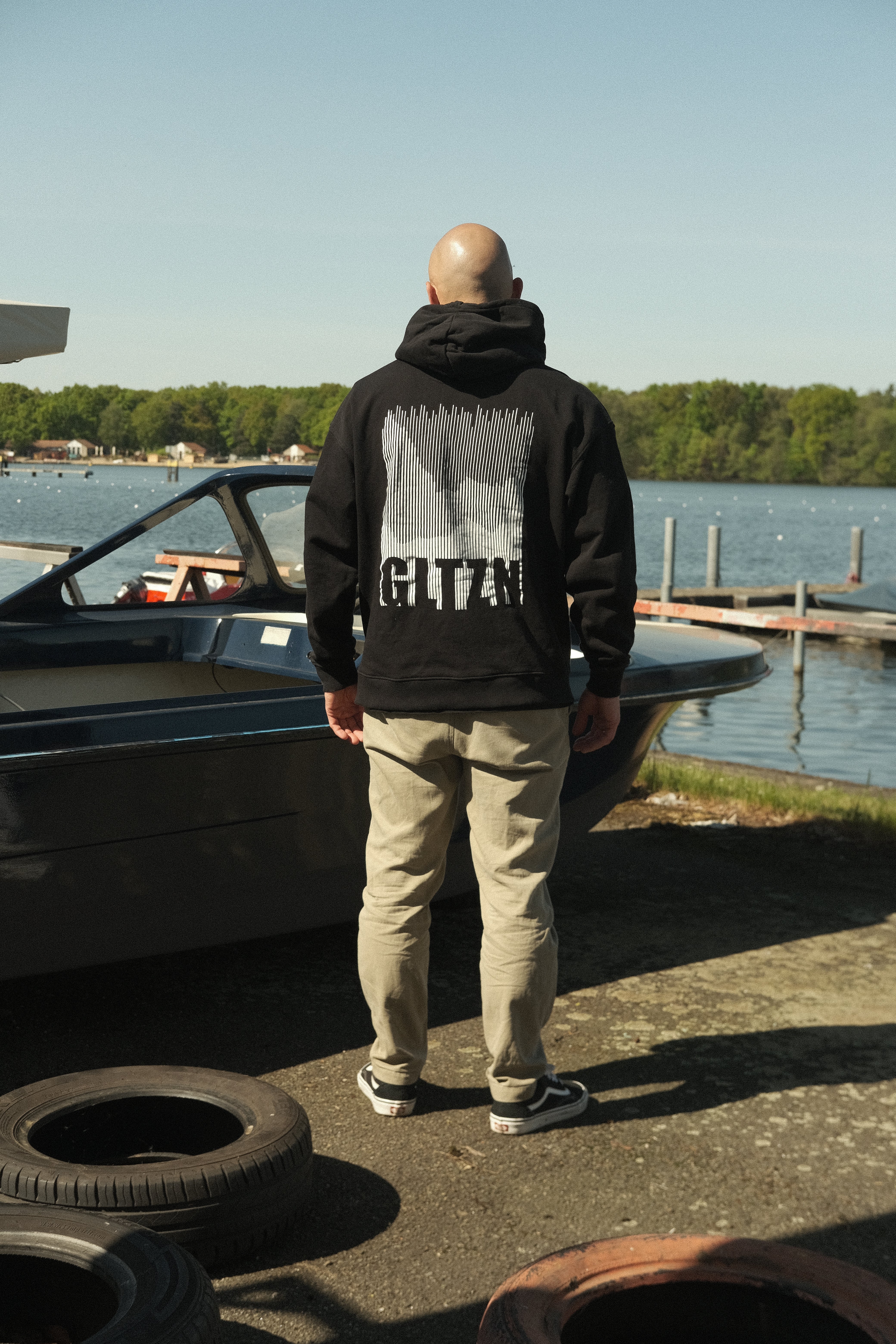 Mann mit Glatze in beiger Hose mit schwarzem GLTZN-Hoodie mit Mountainprint steht vor einem Boot und blickt aufs Wasser