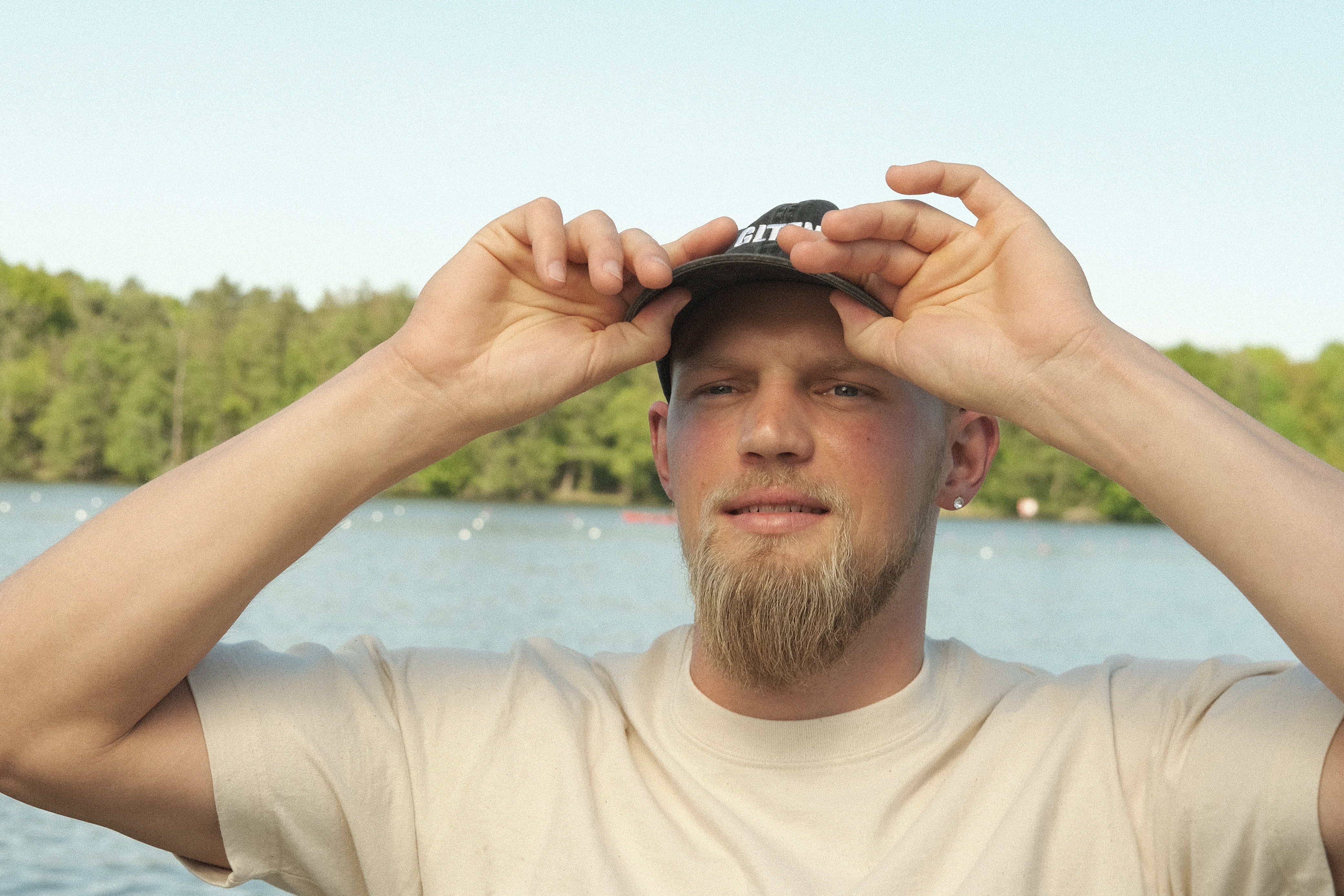 Ein Mann mit Bart trägt eine Kappe am See und hat beide Hände am Kappenschirm