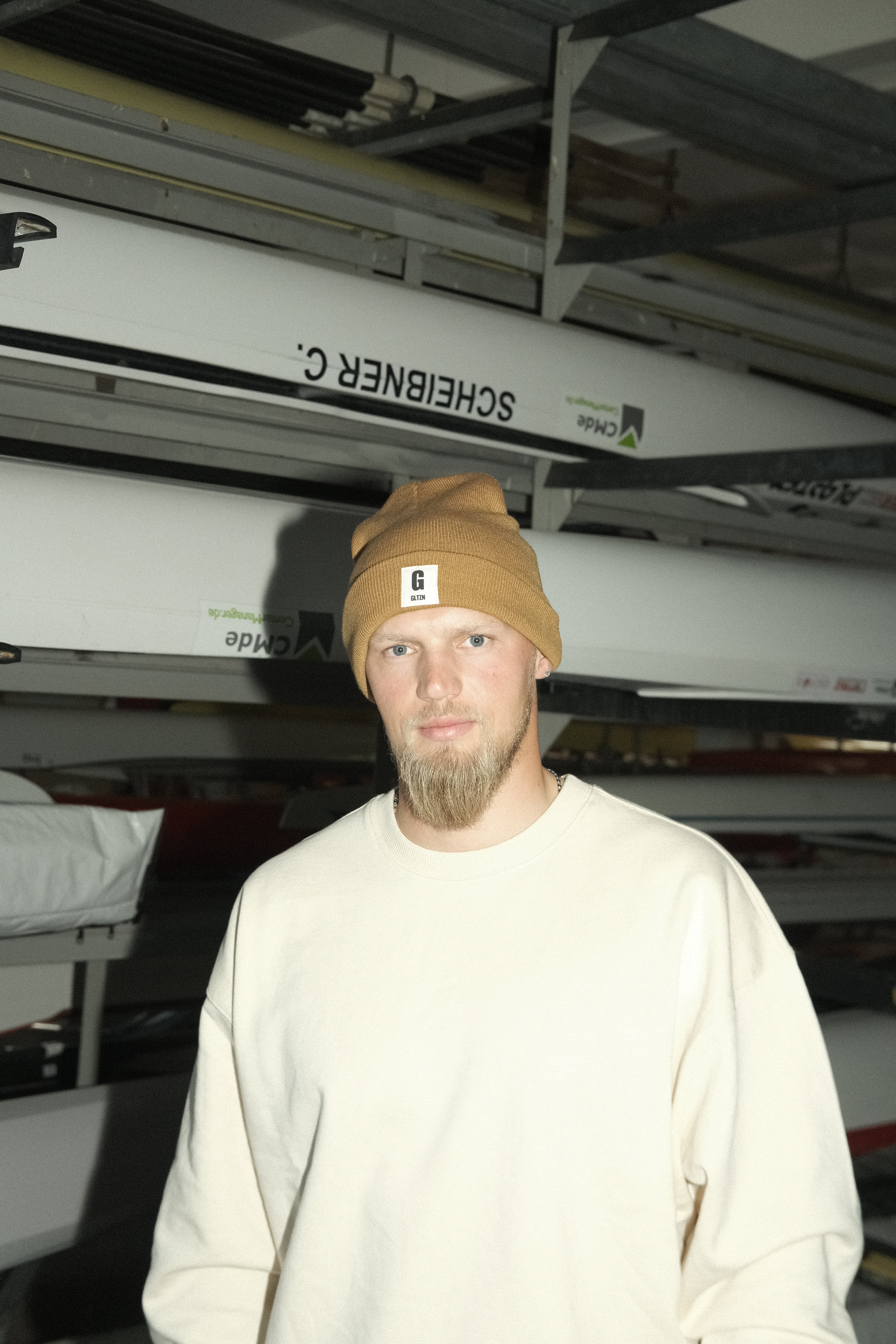Mann mit Bart im beigen Pulli trägt eine braune Beanie mit GLTZN-Logo und steht vor Kanus