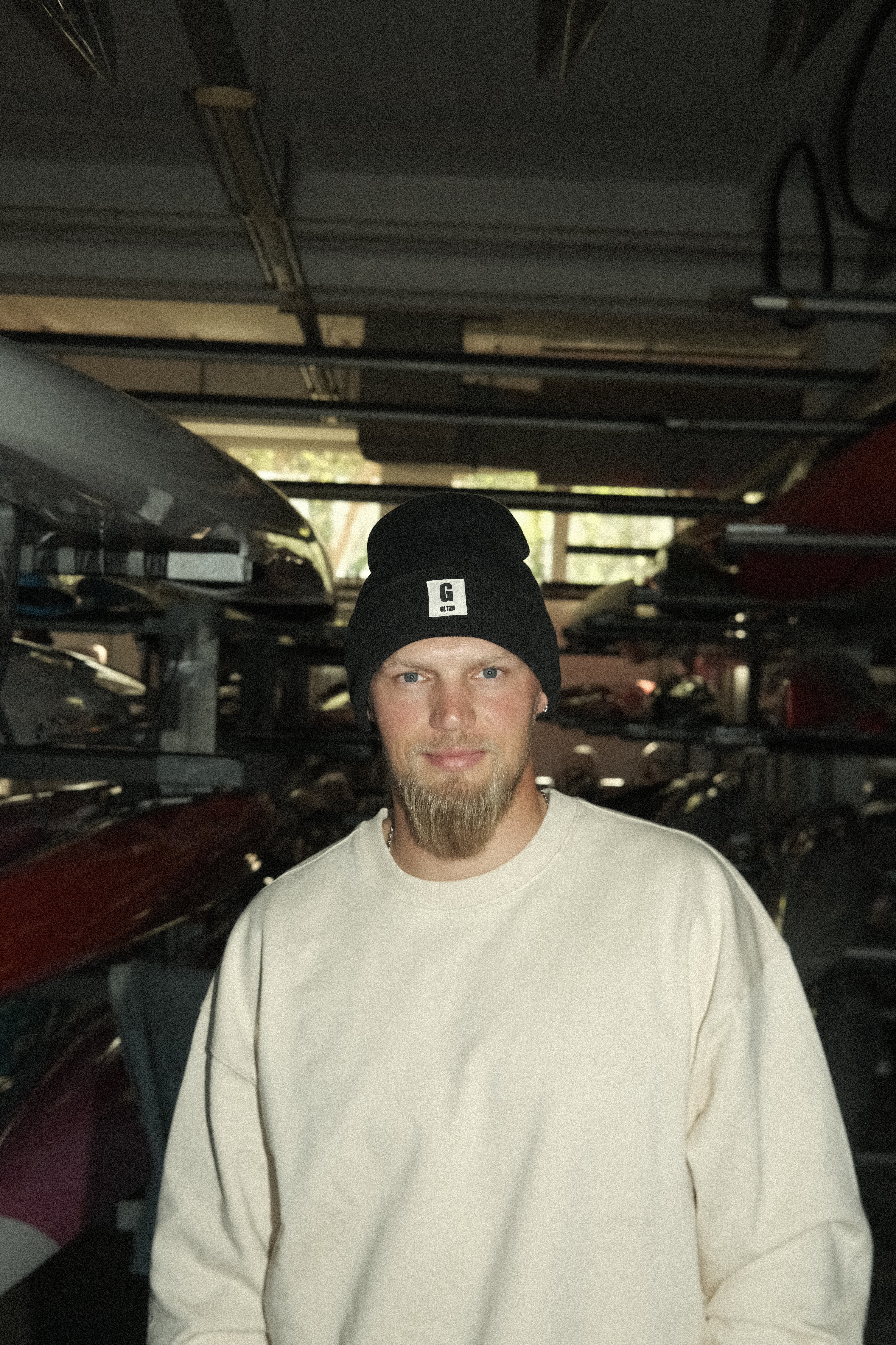 Ein Mann mit Bart im beigen Pulli trägt eine schwarze Beanie mit GLTZN-Logo und steht von mehreren gestapelten Kanus