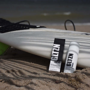 GLTZN MED Sonnencreme LSF 50+ von Phametra im weißen Pumpspender neben der schwarzen Verpackung auf einem Surfbrett im Sand am Strand